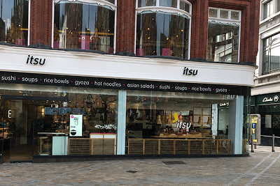 Itsu restaurant storefront with large glass windows displaying menu items like sushi and rice bowls.
