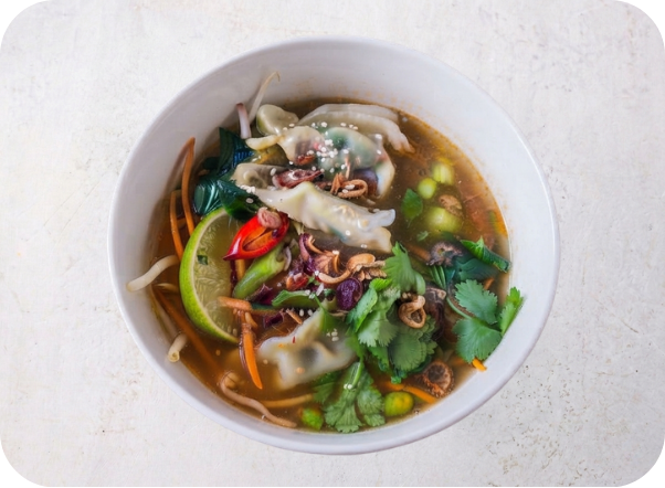 A savory bowl of dumpling soup with vegetables, lime, chili, and cilantro garnish.