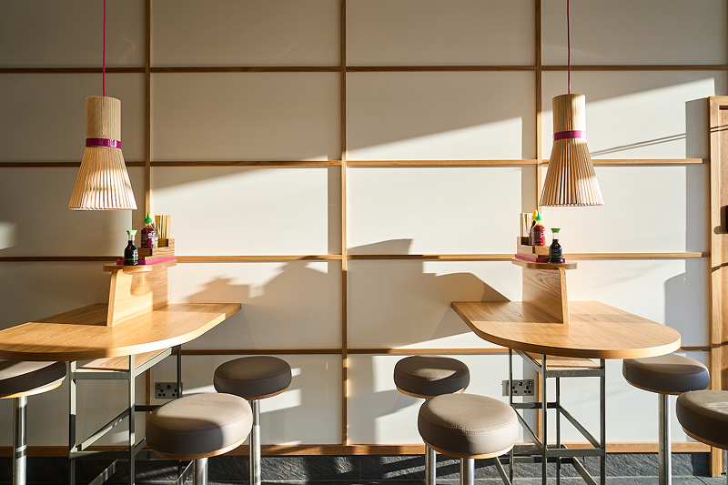 Modern restaurant interior featuring two wooden tables, stools, and pendant lights against a grid wall.