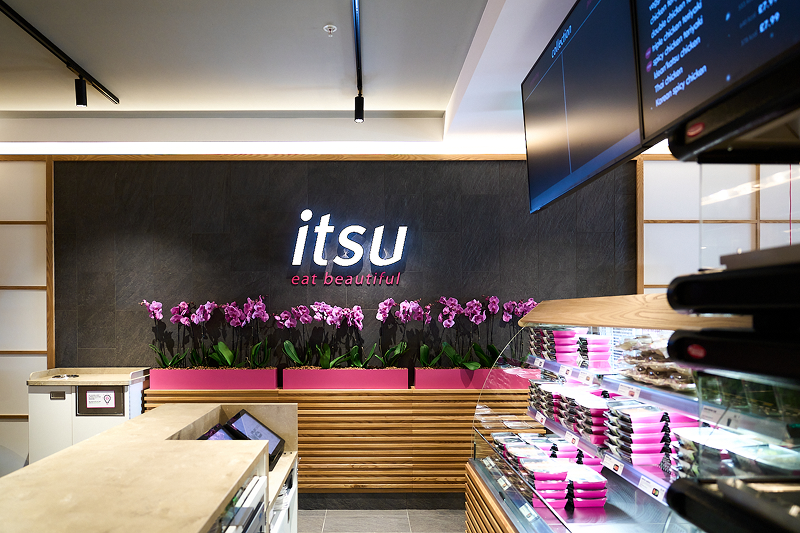 Itsu restaurant interior featuring its illuminated logo, pink orchids, and food display counter.