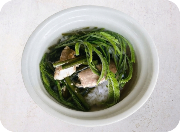 White bowl with green leafy vegetables, pork, and rice, viewed from above.