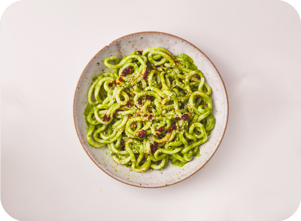 Green pesto pasta in a rustic bowl, sprinkled with red pepper flakes on a light surface.