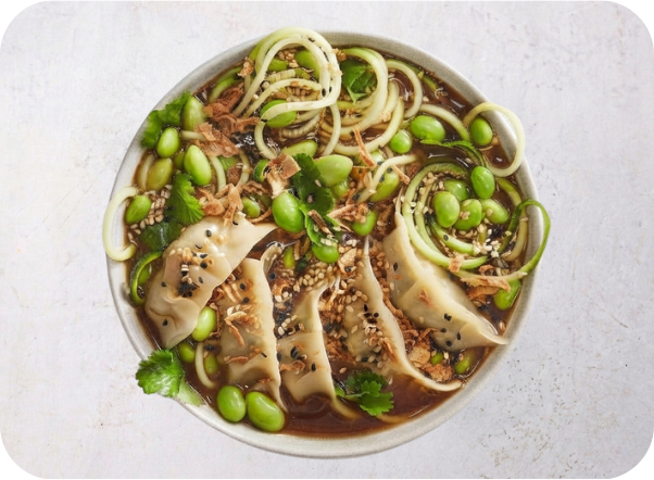 A bowl of savory soup with dumplings, edamame, and zucchini noodles, garnished with sesame seeds.