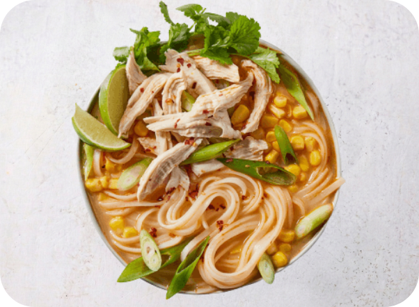 Bowl of chicken noodle soup with corn, green onions, cilantro, and lime wedges.