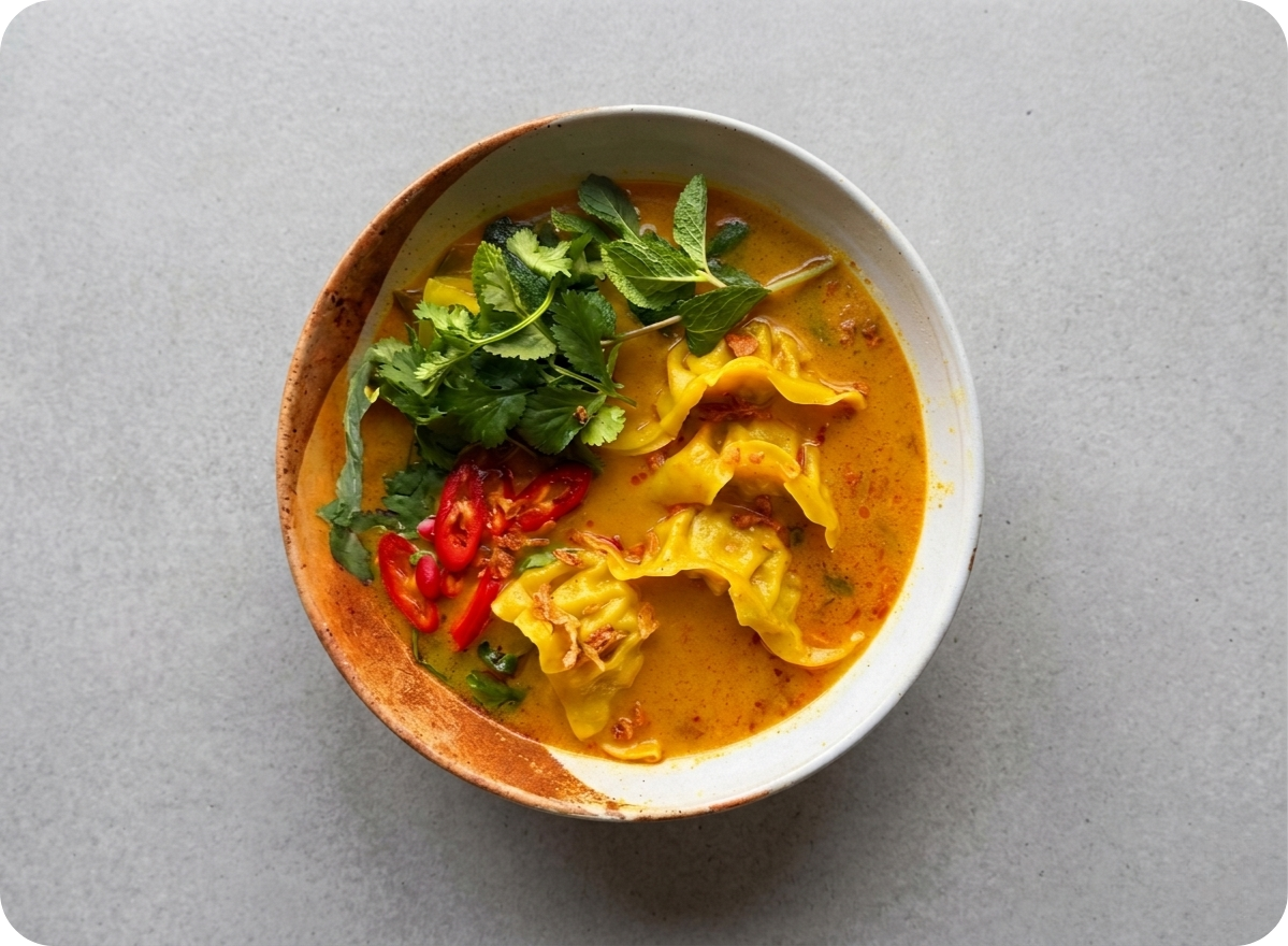 Vibrant orange soup with yellow dumplings, fresh green herbs, and red chili slices.