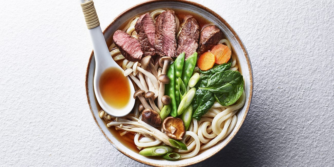 A bowl of ramen with thinly sliced beef, udon noodles, asparagus, mushrooms, and bok choy.