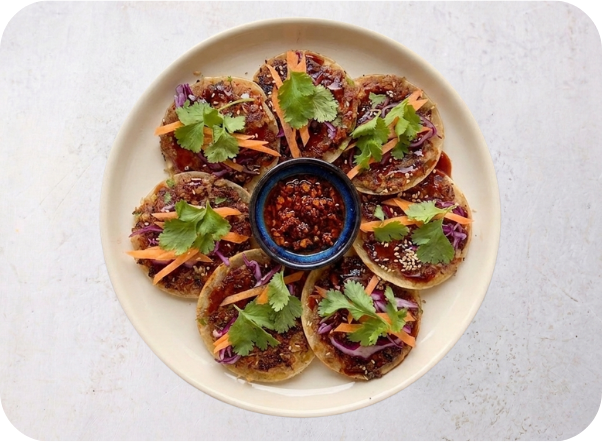 Round appetizers topped with carrots, red cabbage, cilantro, and dark sauce, with chili oil.