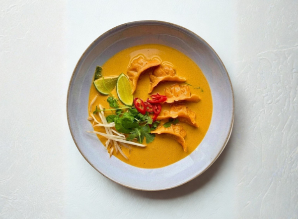 A bowl of vibrant orange curry soup with dumplings, lime, red chilies, and cilantro.