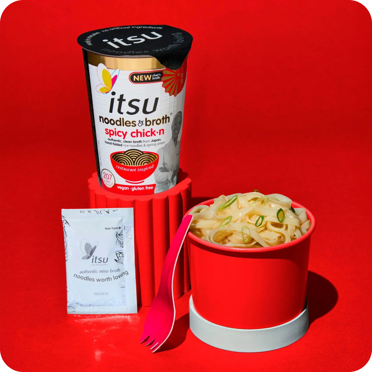 Itsu spicy chick'n noodle cup next to prepared noodles, seasoning packet, and pink spork.