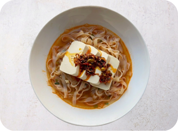 A bowl of flat rice noodles with a block of tofu and chili oil in a flavorful broth.