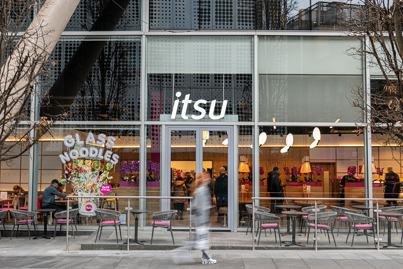 Itsu restaurant storefront with a glass facade, outdoor seating, and blurred people passing by.