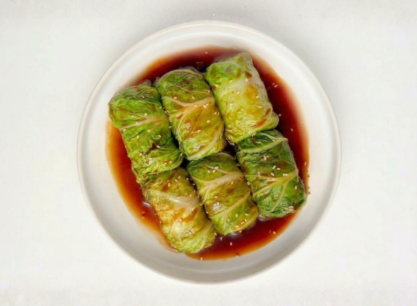 Six green cabbage rolls with sesame seeds in a rich brown sauce on a white plate.