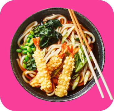 Udon noodle soup with tempura shrimp, green onions, edamame, and seaweed in a dark bowl.