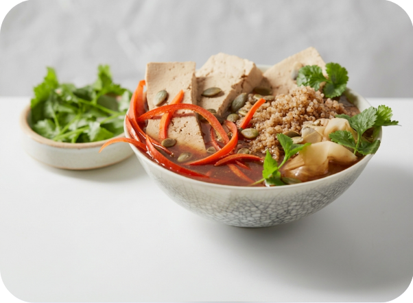 Bowl of savory tofu and vegetable broth with quinoa, carrots, and pumpkin seeds.