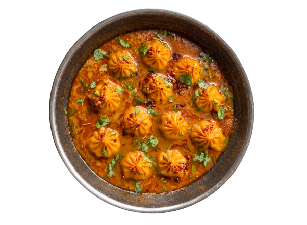 Round metal dish with pleated momos in orange curry, garnished with green cilantro.