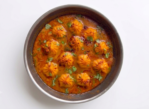 Spicy momos in a rich red gravy, garnished with cilantro, served in a metal pan.
