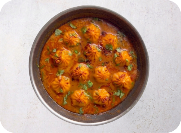 Round metal dish with momos in a rich red curry, garnished with cilantro.
