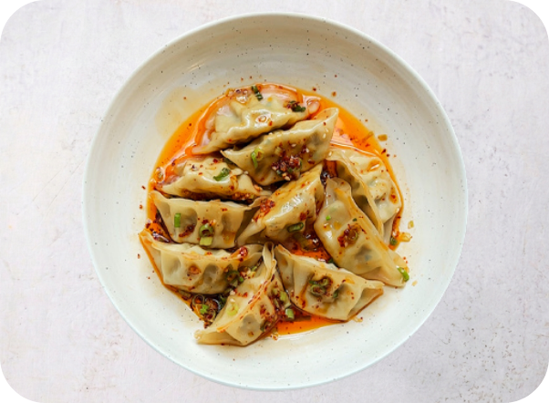 Pan-fried dumplings generously coated in a vibrant chili oil sauce, garnished with chopped green onions.
