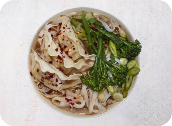 A bowl of noodle soup with dumplings, broccolini, and red chili flakes.