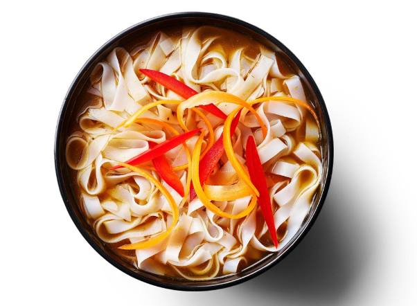 Bowl of flat rice noodle soup with broth, garnished with red and orange pepper strips.