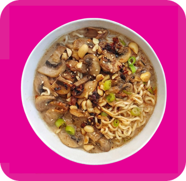 Bowl of ramen noodles with mushrooms, peanuts, and chopped green onions on a pink background.