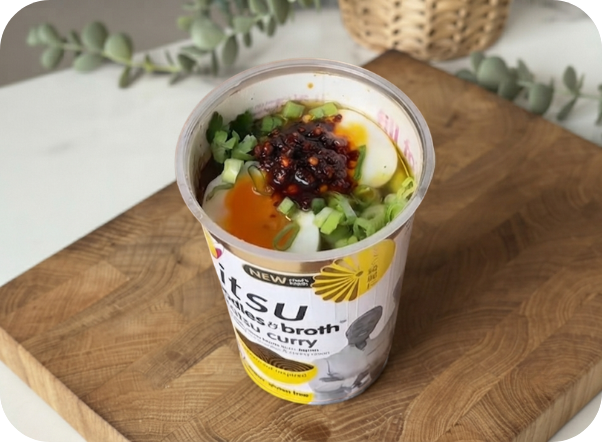 Itsu cup ramen with soft-boiled egg, green onions, and chili oil on a wooden board.
