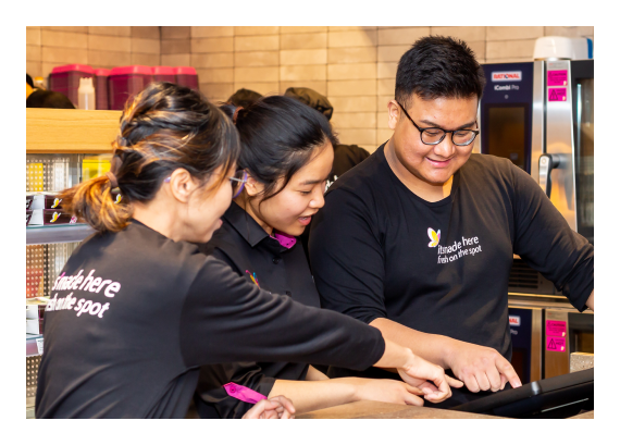 Three smiling employees in black shirts collaborate at a counter, pointing at a digital screen.