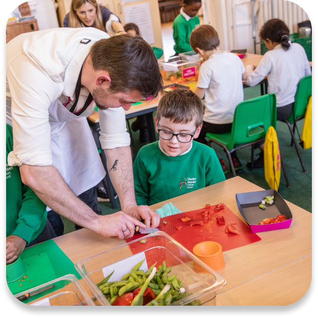 Instructor in apron helps a boy in glasses chop tomatoes during a cooking class.